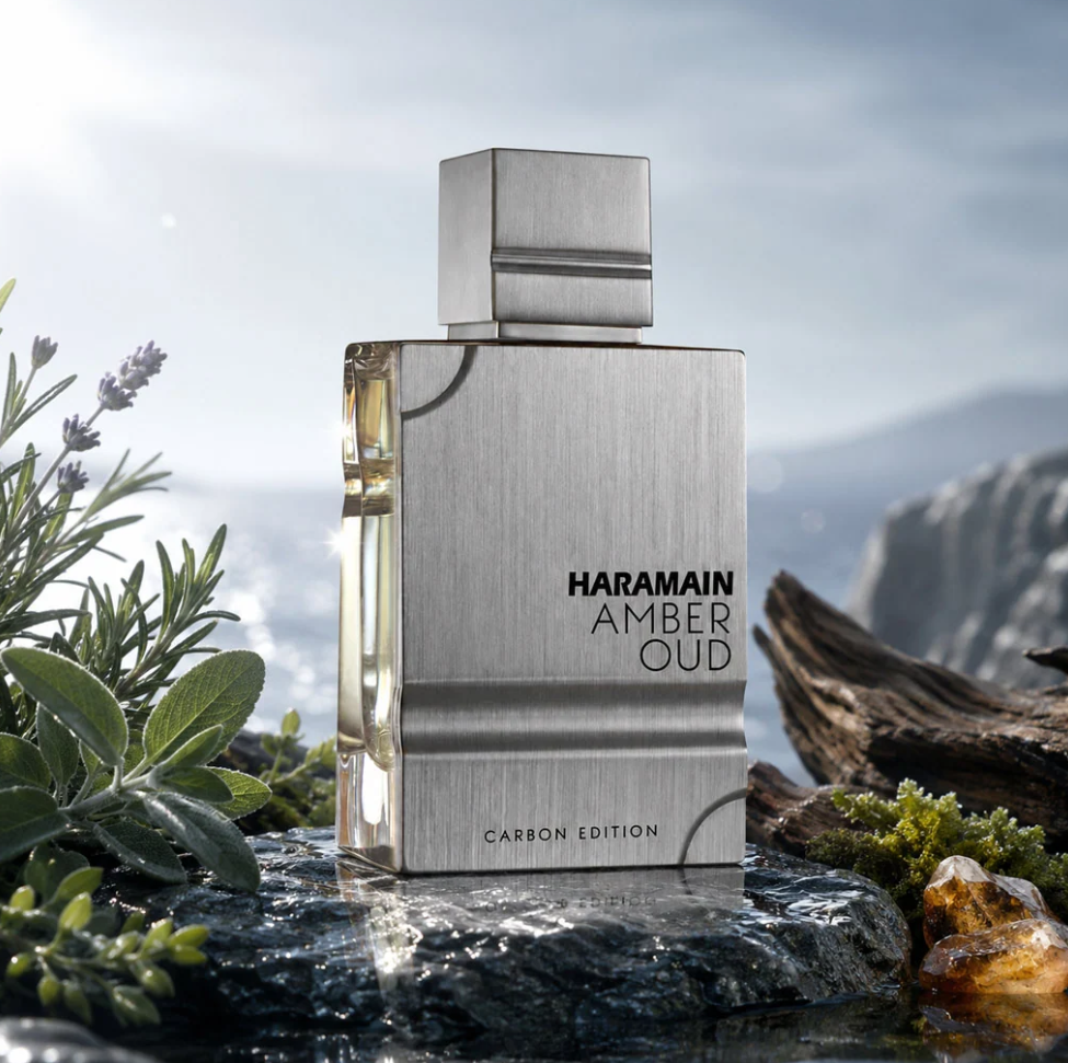 Al Haramain Amber Oud Carbon Edition silver bottle on wet rock beside purple flowers, green herbs, moss and wood, with a misty coast in the background.