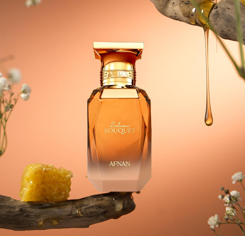 Afnan Delicious Bouquet amber bottle balanced on a wood branch beside a honeycomb-like piece, with dripping golden liquid, small white flowers and an orange background.