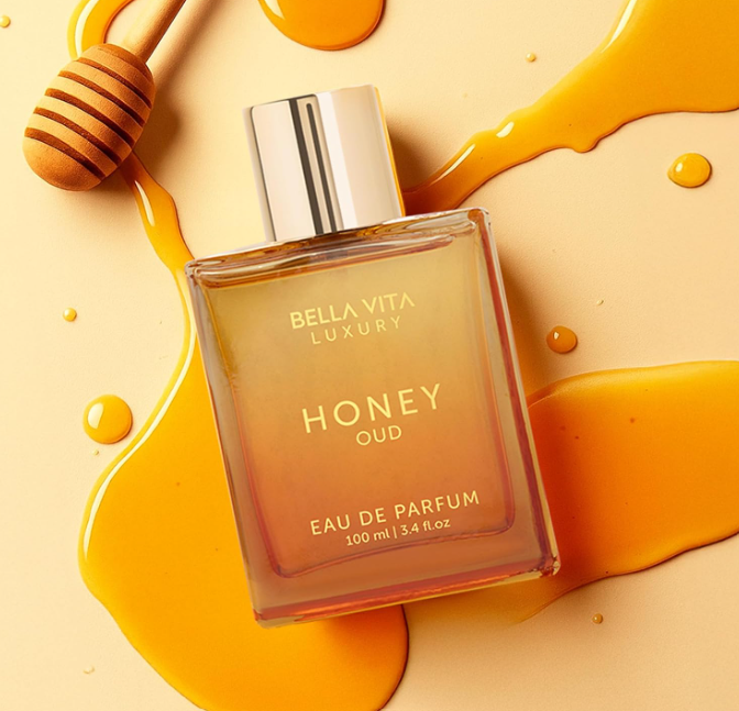 Bellavita Honey Oud amber perfume bottle with a gold cap lying diagonally among golden honey drizzles and a wooden honey dipper on a yellow background.