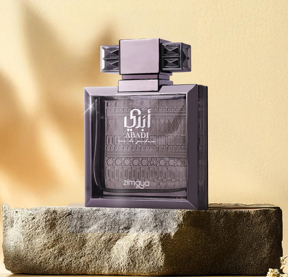 Zimaya Abadi dark-framed patterned bottle with broad geometric cap, standing on a rough stone block against a warm golden wall with soft shadows.