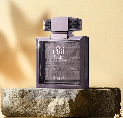 Zimaya Abadi dark-framed patterned bottle with broad geometric cap, standing on a rough stone block against a warm golden wall with soft shadows.
