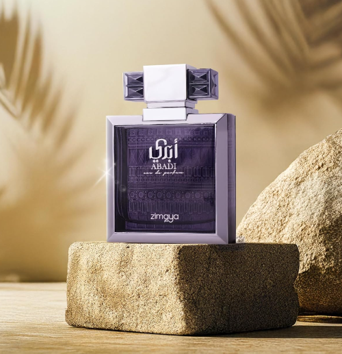 Zimaya Abadi Opulent purple bottle with metallic frame and wide geometric cap on a tan stone block, beside a larger rock against a warm beige background with leaf shadows.
