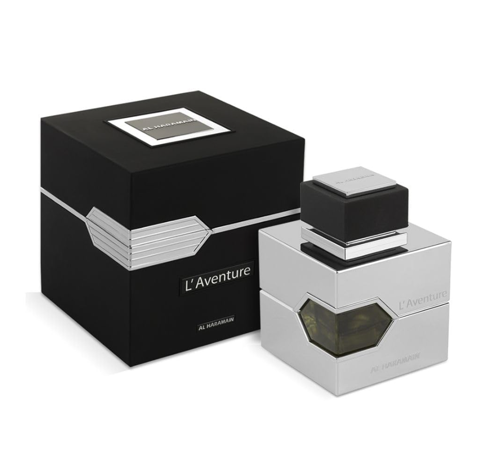 Al Haramain L'Aventure silver square perfume bottle with an amber glass window and black cap beside a black box with silver geometric detailing on white.