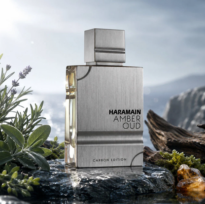 Al Haramain Amber Oud Carbon Edition silver bottle on wet rock beside purple flowers, green herbs, moss and wood, with a misty coast in the background.