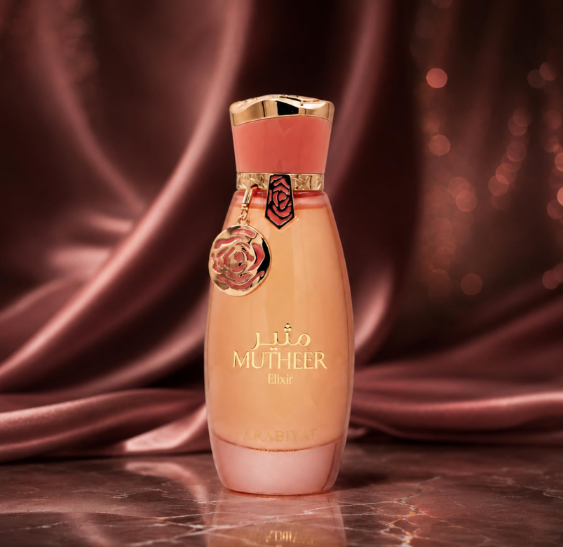 Arabiyat Prestige Mutheer Elixir peach bottle with coral-and-gold cap and hanging charm, on a reflective brown surface in front of draped burgundy satin.