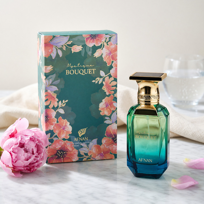 Afnan Mystique Bouquet green-and-blue bottle and floral box on pale marble, beside a pink flower and petals, with cream fabric and clear glass in the background.
