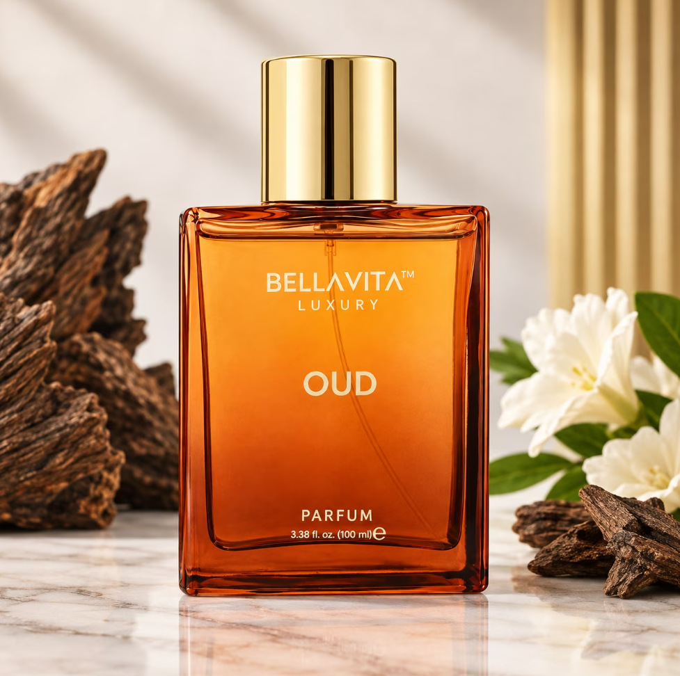 Bellavita Oud Parfum amber bottle with a gold cap on a pale marble surface, framed by dark wood pieces, white flowers and a gold-toned background accent.