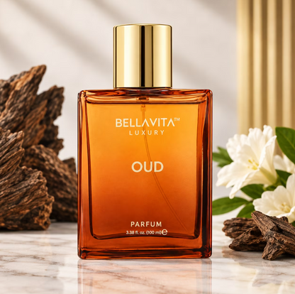 Bellavita Oud Parfum amber bottle with a gold cap on a pale marble surface, framed by dark wood pieces, white flowers and a gold-toned background accent.