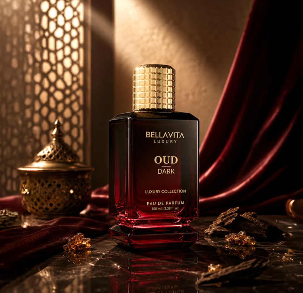 Bellavita Oud Dark burgundy bottle with a textured gold cap on reflective stone beside an ornate metal vessel, dark red fabric, wood and amber-colored pieces.