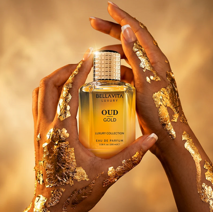 Hands decorated with gold leaf holding a Bellavita Oud Gold perfume bottle with golden liquid and a textured gold cap against a warm gold background.