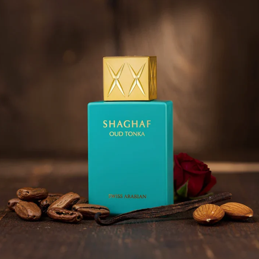 Swiss Arabian Shaghaf Oud Tonka turquoise bottle with gold cap on a wooden surface, beside almonds, dark seeds and dark pods and a red rose on a brown background.