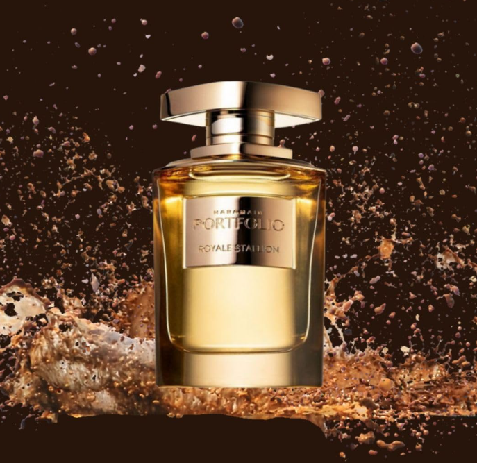 Al Haramain Portfolio Royale Stallion gold-toned perfume bottle and broad gold cap surrounded by a splash of amber-brown liquid against a dark brown background.