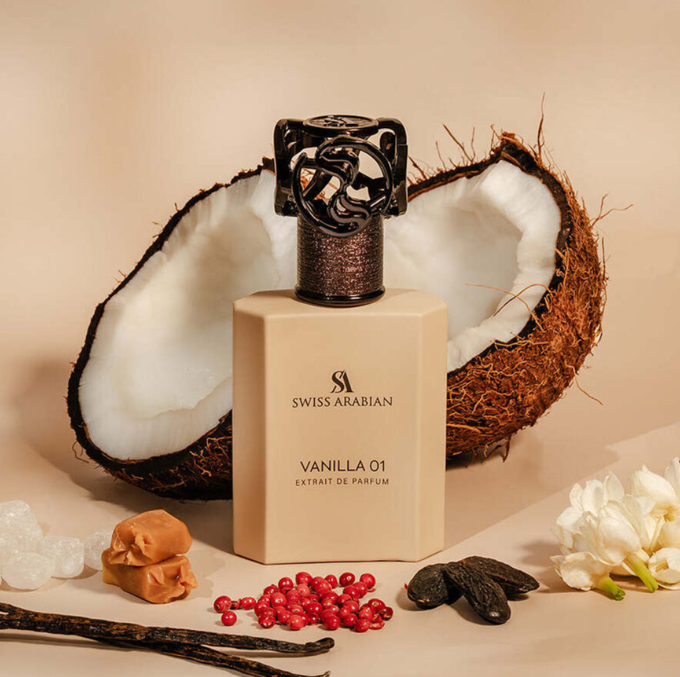 Swiss Arabian Vanilla 01 beige bottle on a tan background with split coconut, white flowers, red berries, dark pods, amber-colored pieces and a small brown sweet-shaped prop.