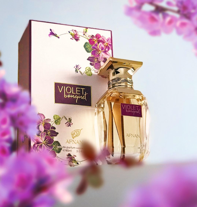 Afnan Violet Bouquet clear bottle with gold cap beside its cream floral box, framed by blurred purple and pink flowers against a pale blue background.