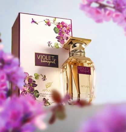 Afnan Violet Bouquet clear bottle with gold cap beside its cream floral box, framed by blurred purple and pink flowers against a pale blue background.