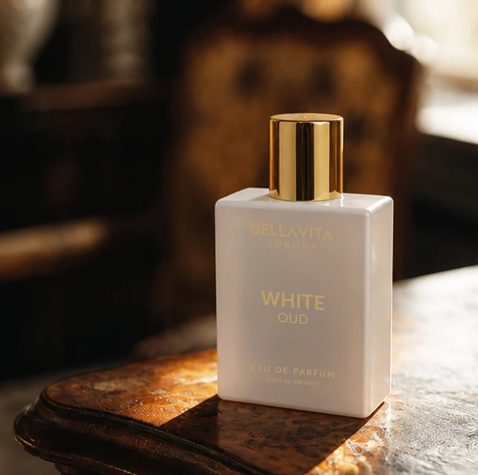 Bellavita White Oud white perfume bottle with a gold cap on a sunlit wooden tabletop, with a softly blurred brown chair in the background.